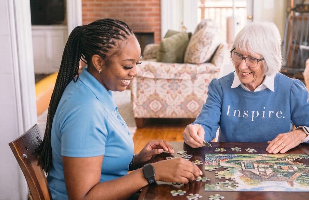 CaraVita caregiver in a light blue polo working on a jigsaw puzzle with an older woman at her dining table